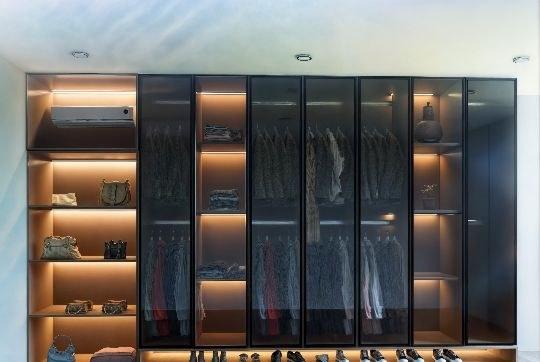 Full-wall wardrobe design with smoked glass doors, warm LED shelf lighting and an open shoe display